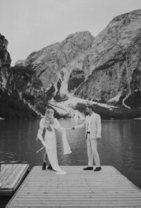 A bride and groom hold hands in their wedding attire while looking out over Lago Di Braies in the Italian Dolomites