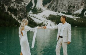 A bride and groom hold hands in their wedding attire while looking out over Lago Di Braies in the Italian Dolomites