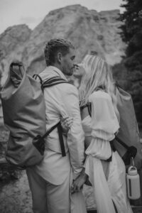 A black and white photo of an elopement couple in wedding attire standing at Lago Di Braies with hiking bags on. 