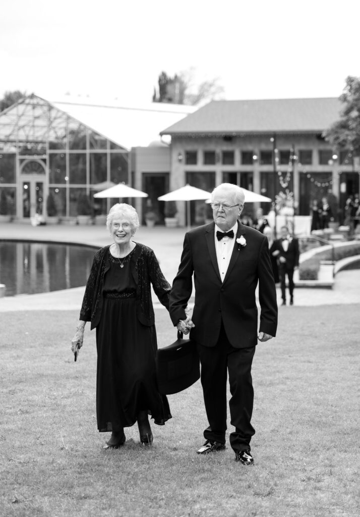 Black and white photo of an elderly couple walking beside the reflection pond at La Caille wedding venue in Utah during a wedding celebration