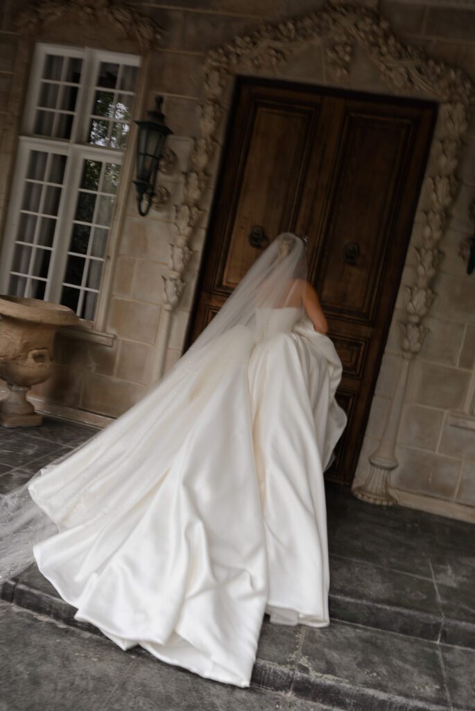 A bride in a Nicole Felicia ballgown and floor length veil walks into the doors of a French chateau at La Caille Wedding Venue in Utah