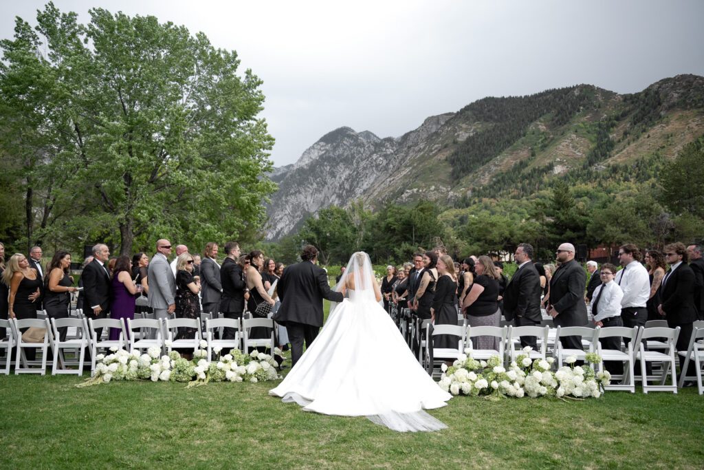 Outdoor wedding ceremony at La Caille wedding venue in Utah with mountain backdrop, featuring a bride walking down the aisle toward seated guests
