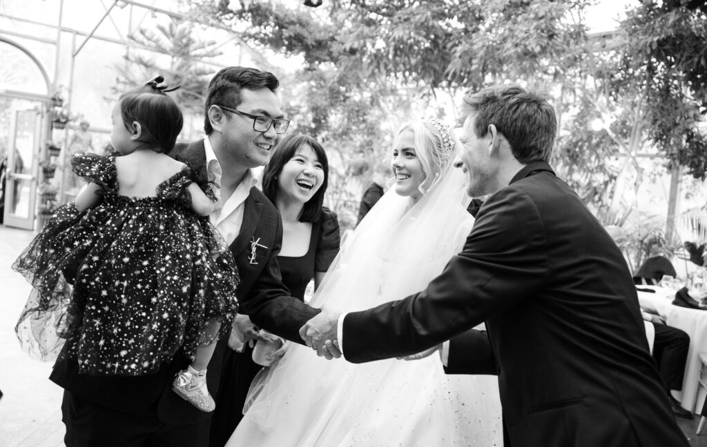 Candid black and white photo of wedding guests sharing laughter inside the La Caille greenhouse during a wedding celebration in Utah